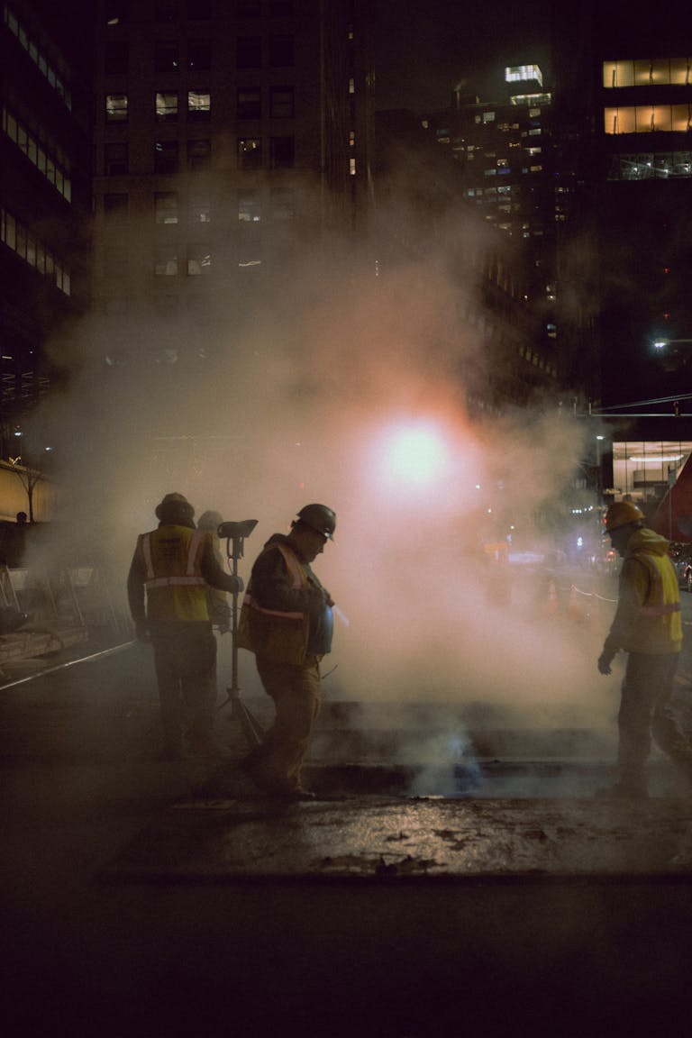 Men working on city streets at night amidst urban lights and steam.