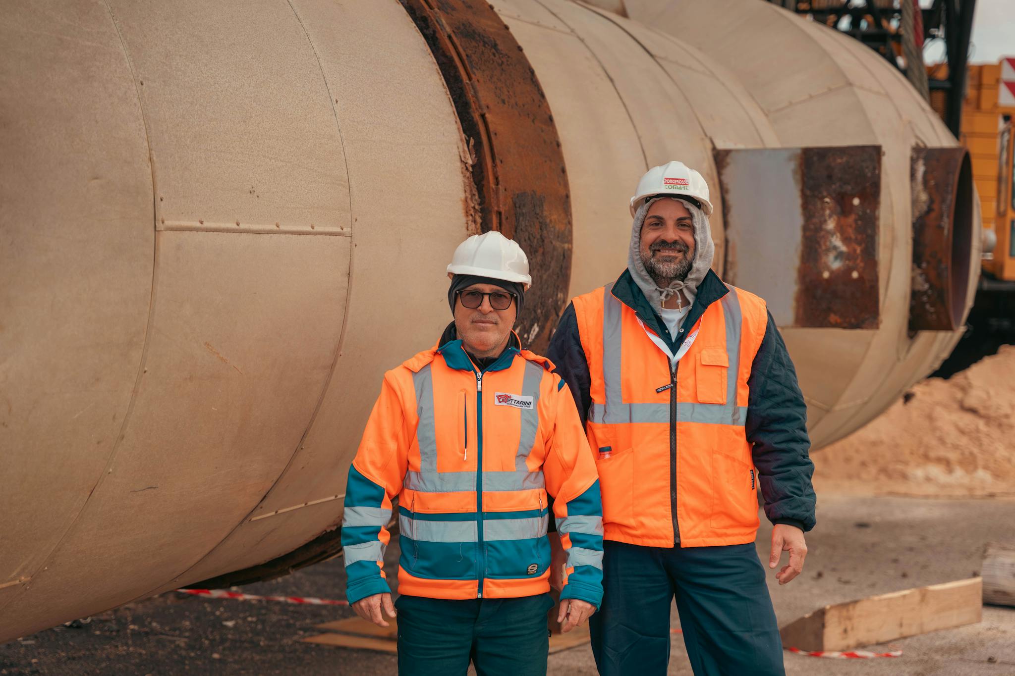 Two construction workers in safety gear standing by industrial machinery on a construction site.